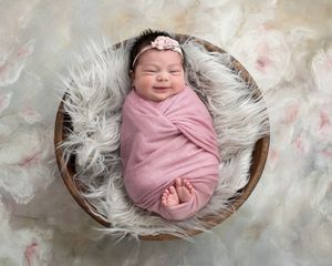 Smiling newborn baby swaddled in pink wrap with floral headband, resting in a wooden bowl on fluffy white fur.