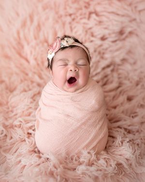 Adorable newborn baby girl swaddled in pink wrap, wearing floral headband, yawning on fluffy pink fur backdrop.