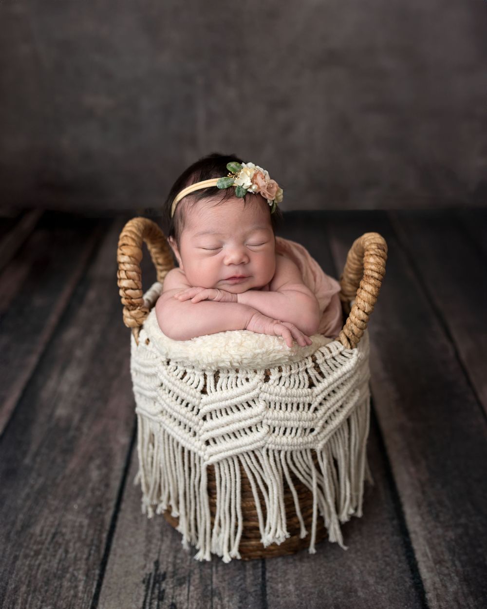 Newborn baby girl sleeping peacefully in a macrame basket with a floral headband on a rustic wood floor.