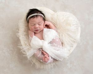 Sleeping newborn baby girl wrapped in white bow in heart-shaped fluffy basket with diamond headband.