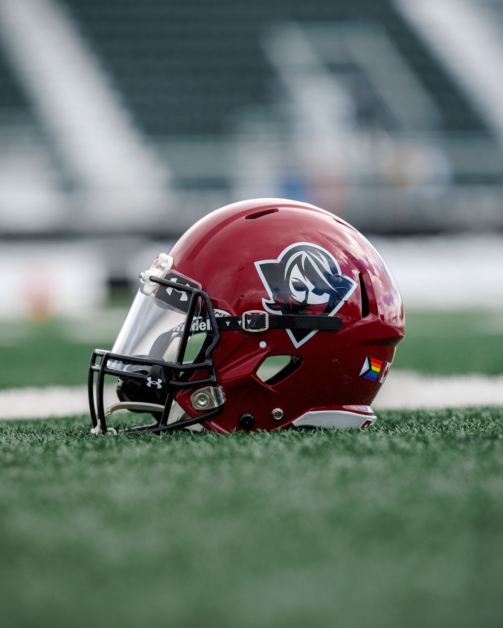 Red football helmet with logo resting on green turf in an empty stadium.