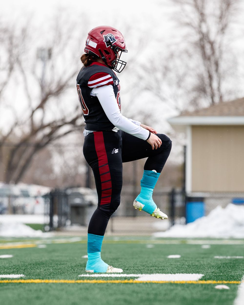 Female football player in red helmet and black uniform stretching on outdoor turf field.