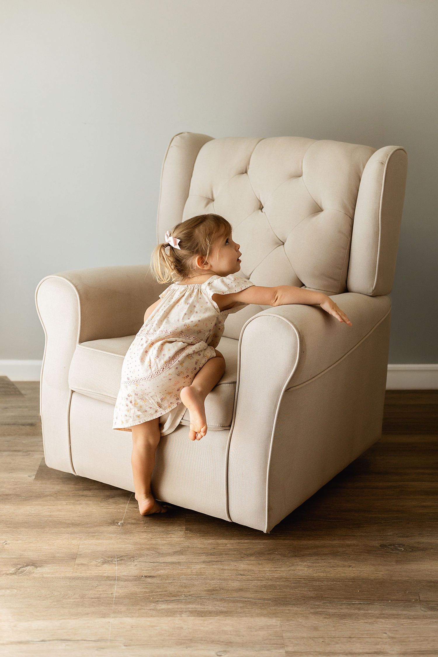 A toddler girl in a floral dress climbs onto a large beige tufted armchair on a hardwood floor.