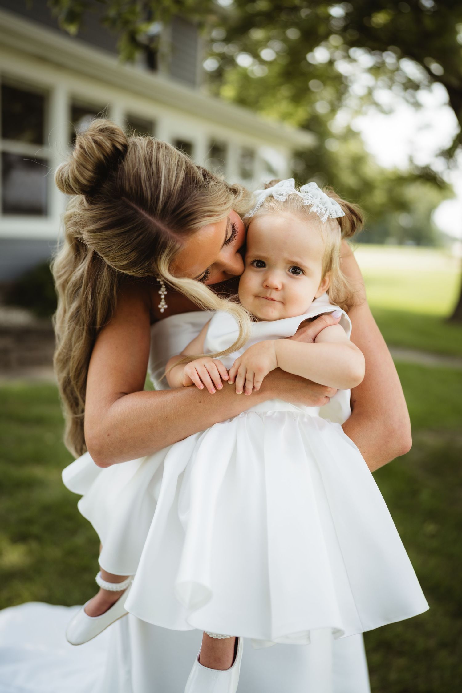 Bride in white gown helps toddler flower girl walk on green lawn outside a gray house on a wedding day.