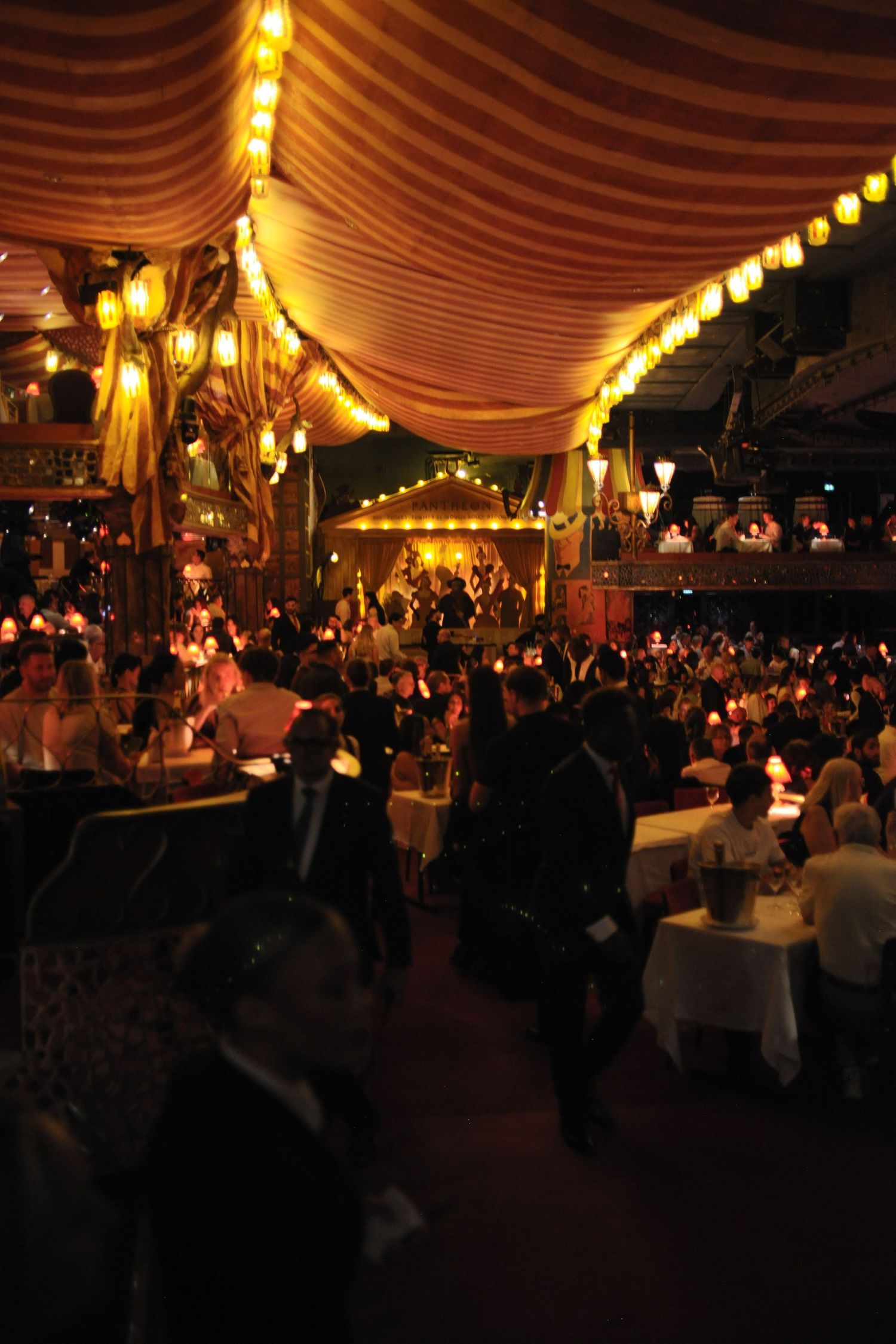 Woman seated at candlelit table in dimly lit theater venue with red curtain stage and audience dining in background.