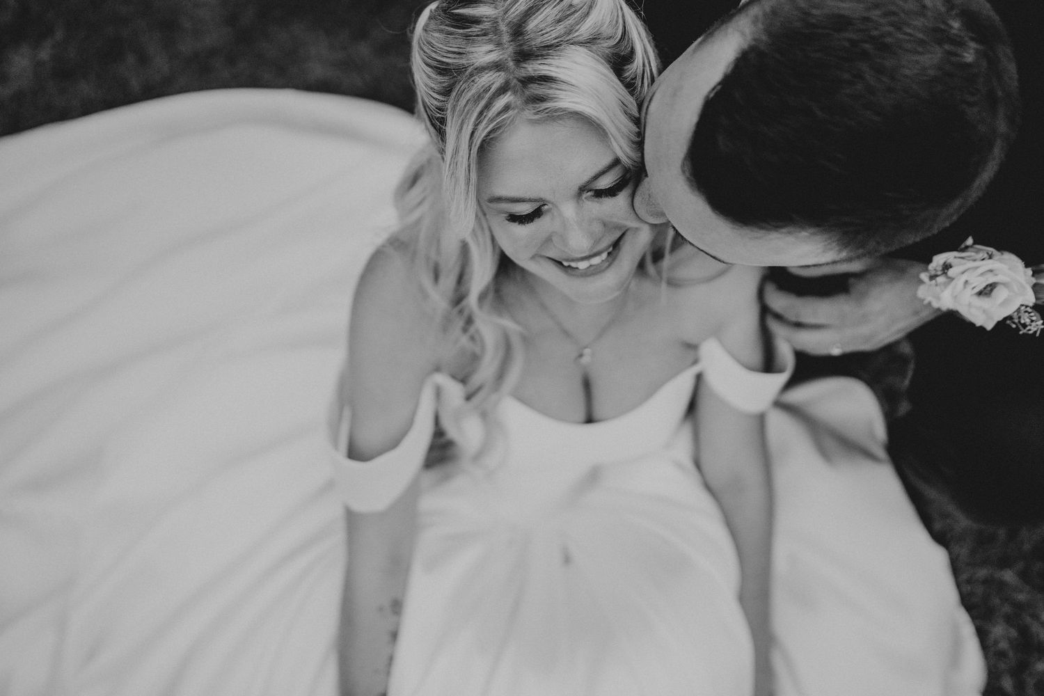 Black and white wedding photo of smiling bride in strapless gown as groom tenderly kisses her cheek.