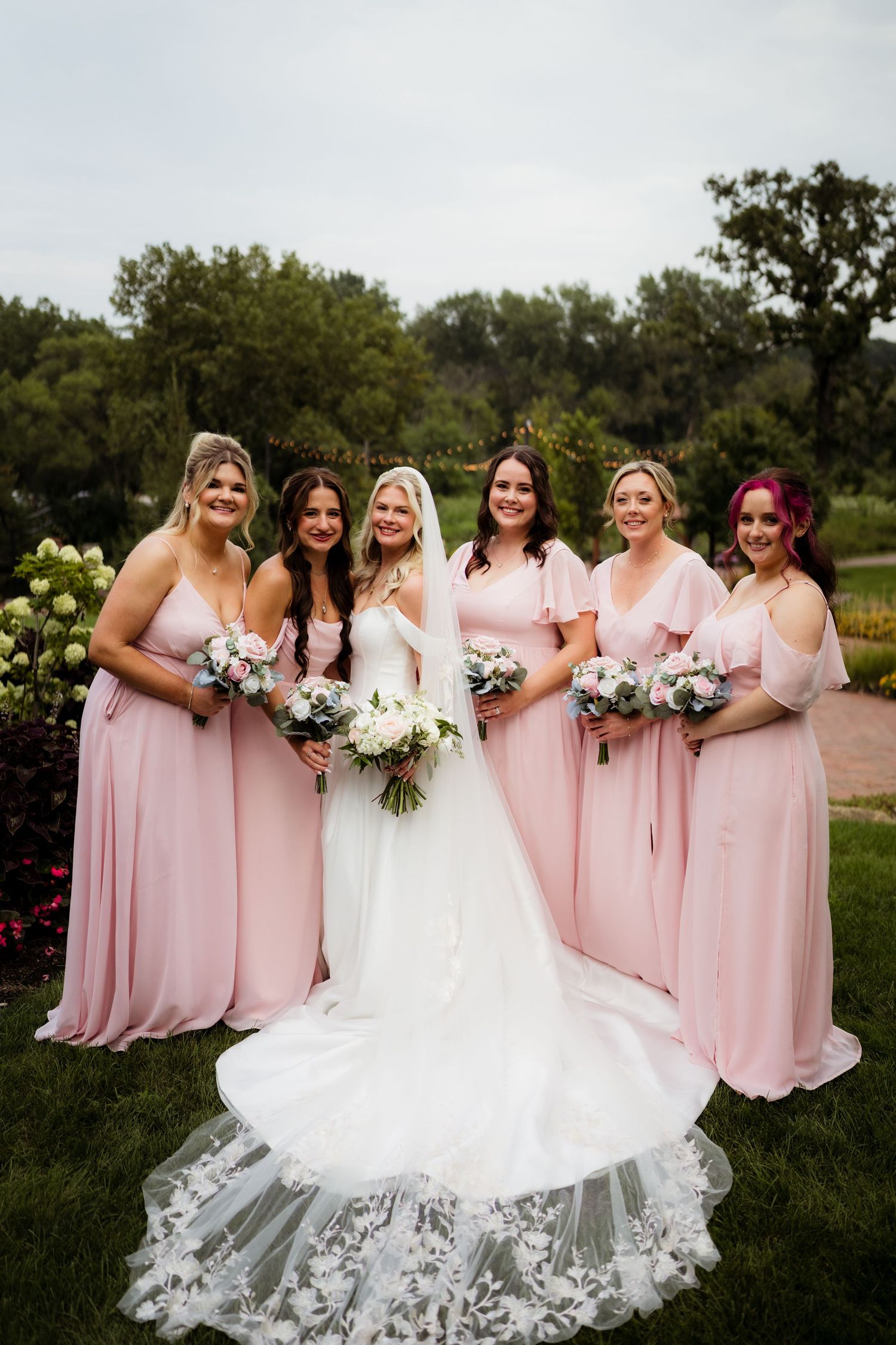 Bride in white gown with five bridesmaids in pink dresses holding bouquets, posing outdoors in a garden setting.