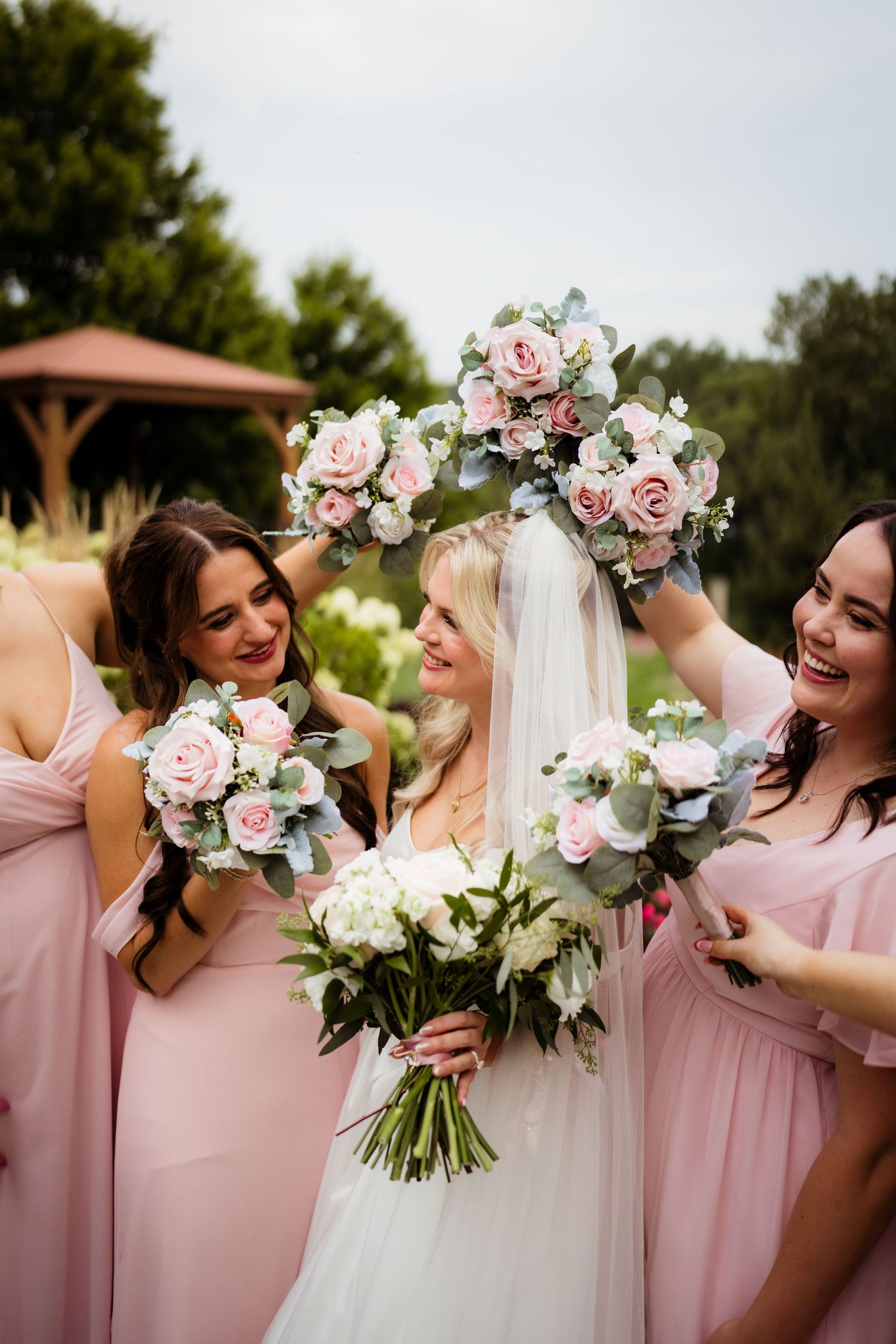 Bride in white gown holds bouquet surrounded by bridesmaids in pink dresses raising floral arrangements outdoors.