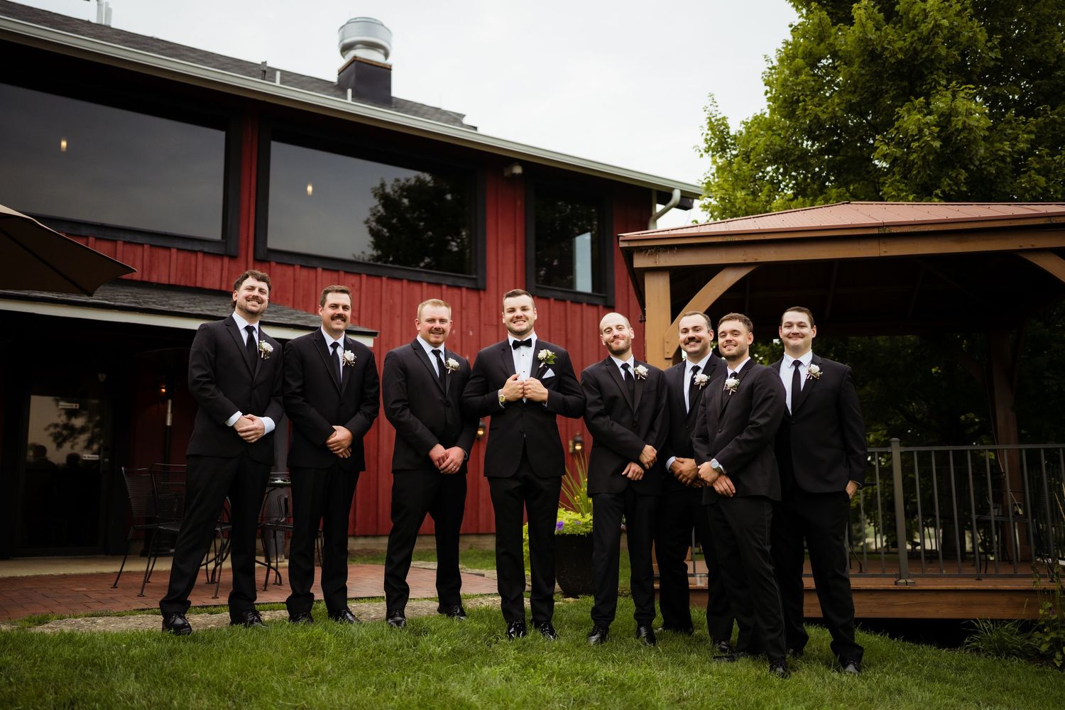 Groom and groomsmen in black suits posing outdoors in front of a red barn venue on a wedding day.