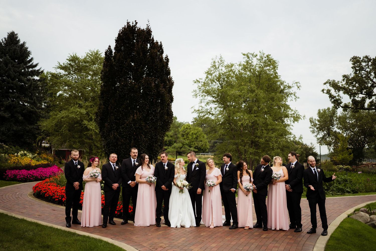 Wedding party poses outdoors on brick path surrounded by lush green trees and colorful flowers.