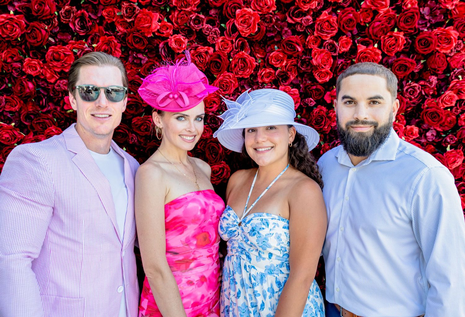 Four elegantly dressed guests pose before a red rose wall, wearing pastel suits and colorful fascinators at a formal event.