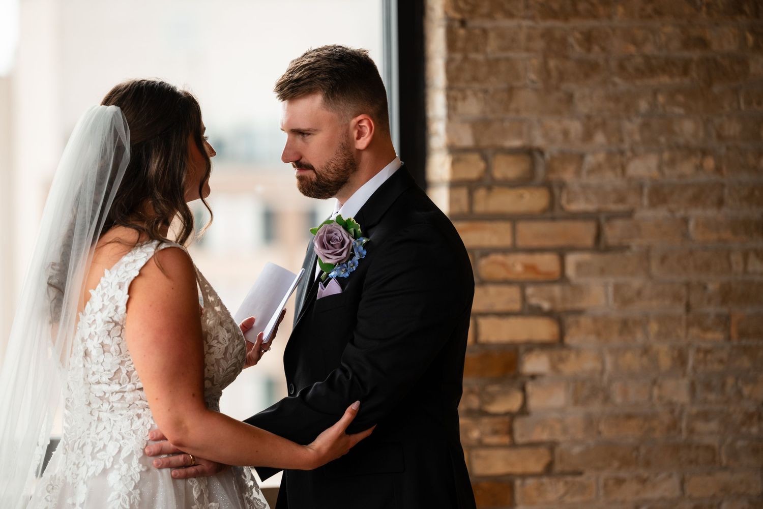 Bride and groom share an intimate moment exchanging vows near a brick wall window during their wedding ceremony.