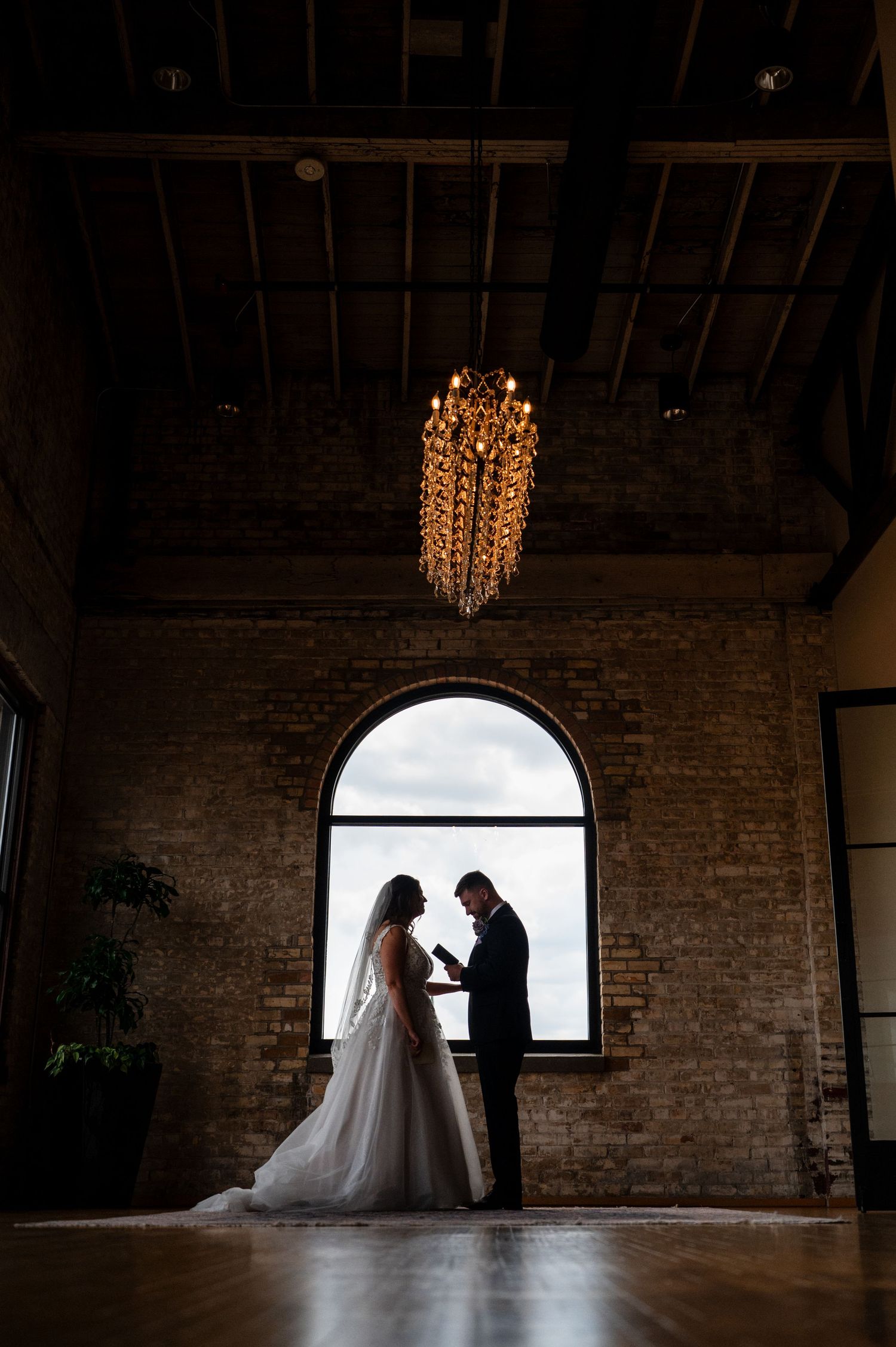 Silhouette of bride and groom exchanging vows by arched window in rustic brick venue with chandelier.