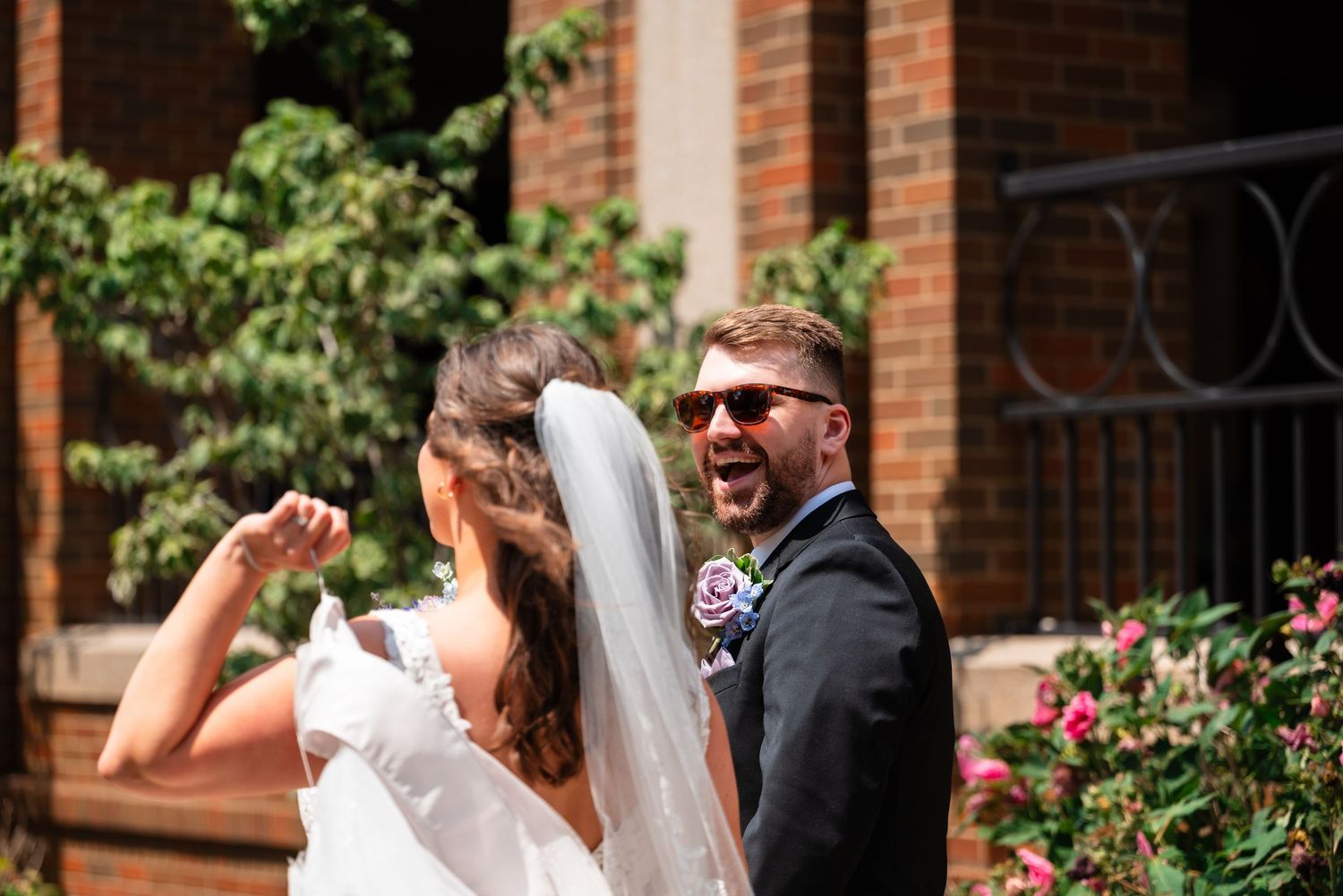 Excited groom in sunglasses reacts to seeing bride in white wedding dress and veil outside brick building.