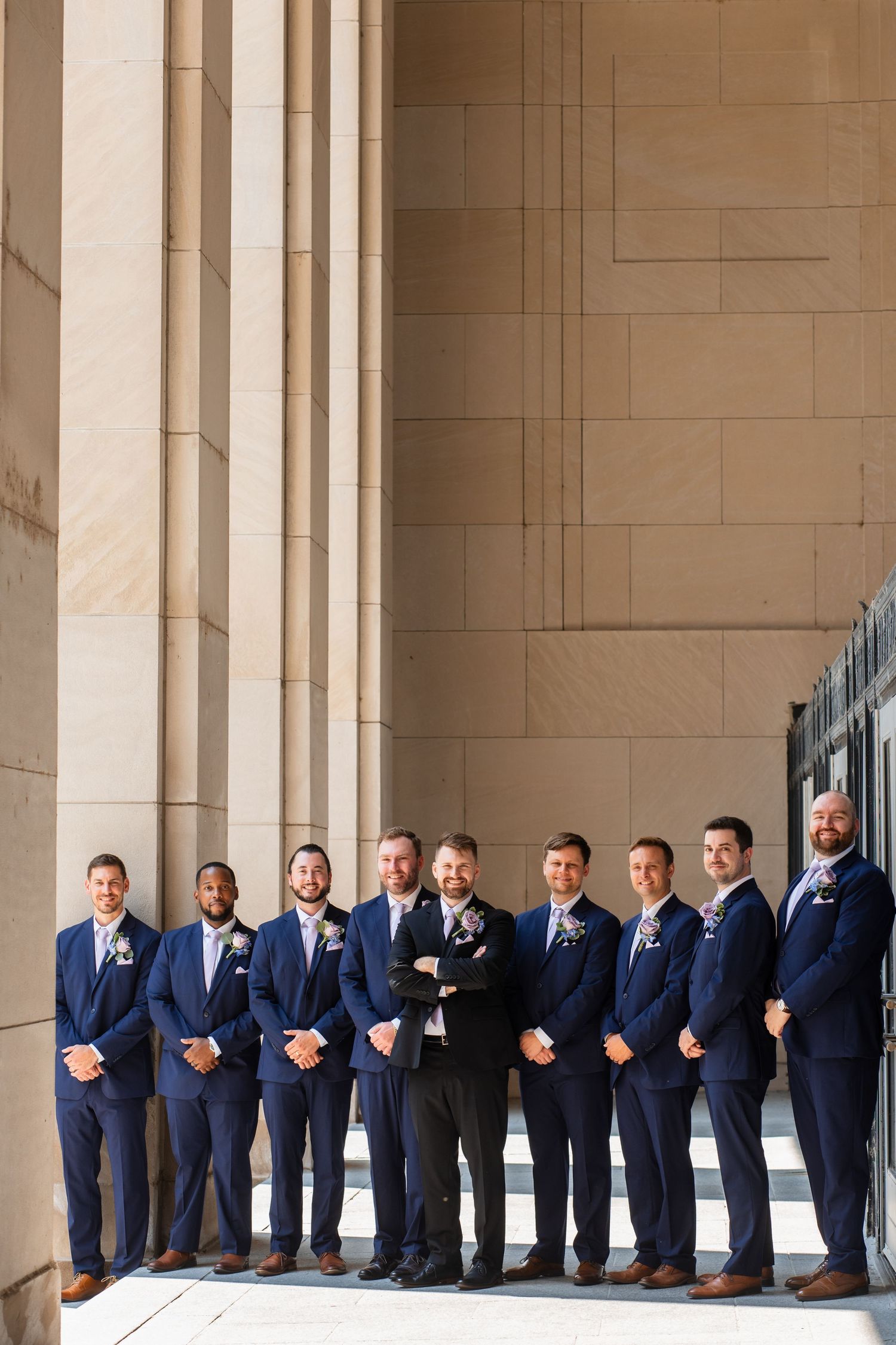 Groom in black suit stands with eight groomsmen in navy blue suits and boutonnieres beside stone columns.