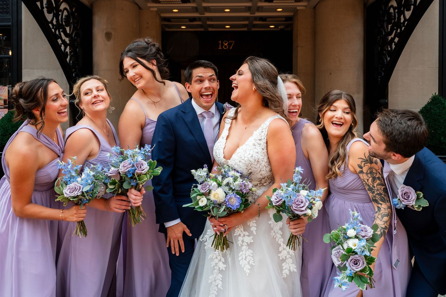 Bride and groom laugh joyfully with bridesmaids in lavender dresses holding blue bouquets outside elegant venue.