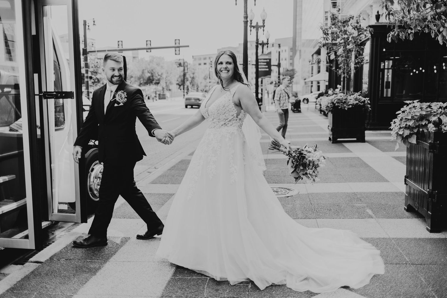 Bride and groom walk hand-in-hand on city street, bride in flowing white gown holding bouquet, black and white photo.