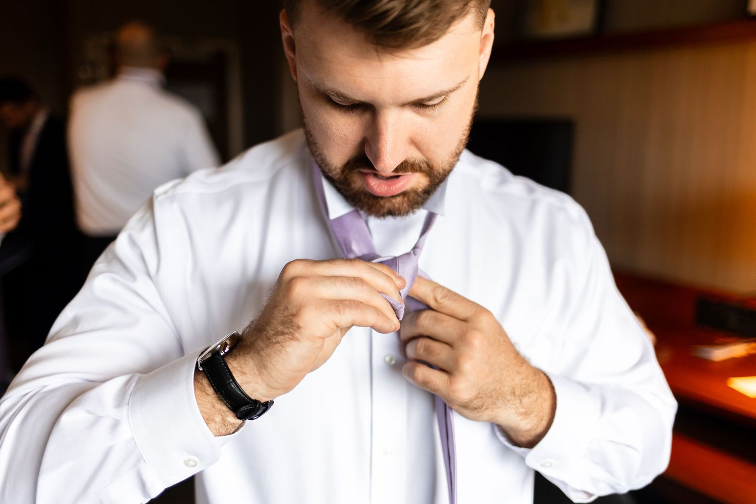 Man in white dress shirt adjusting his purple tie, preparing for a formal event or wedding ceremony.