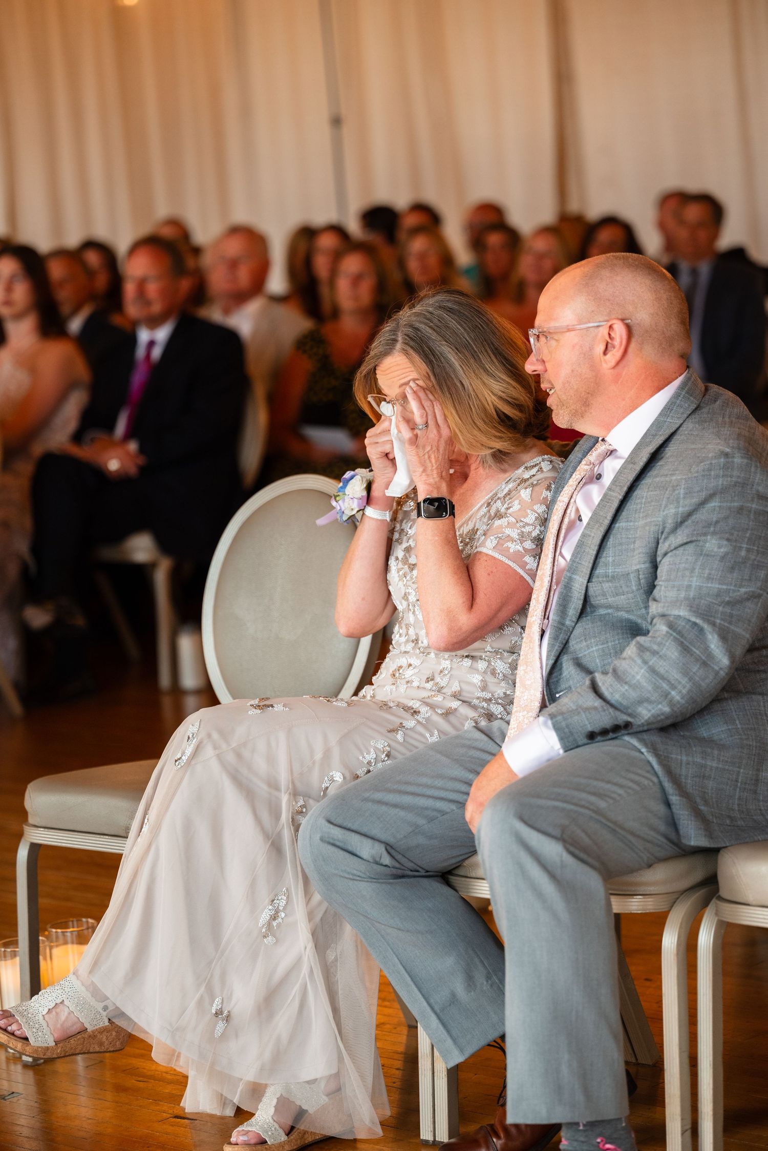 Emotional mother wiping tears during wedding ceremony seated beside man in gray suit at elegant venue.
