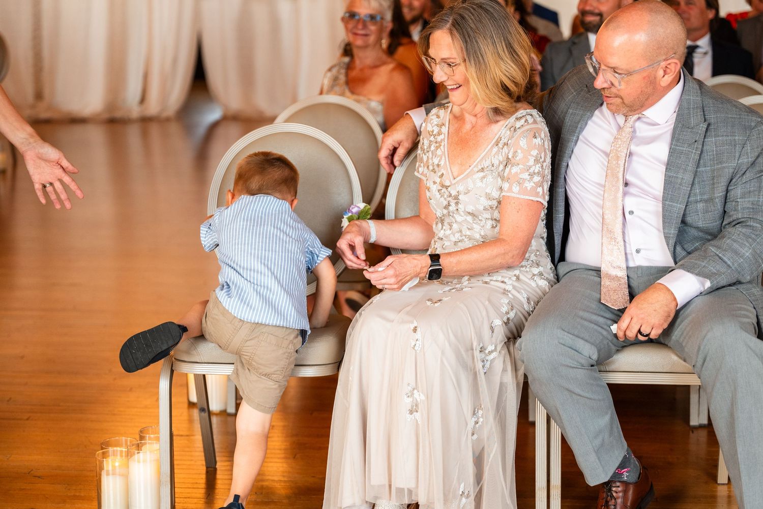 A young boy in a blue shirt climbs on chairs near smiling adults at a wedding ceremony.