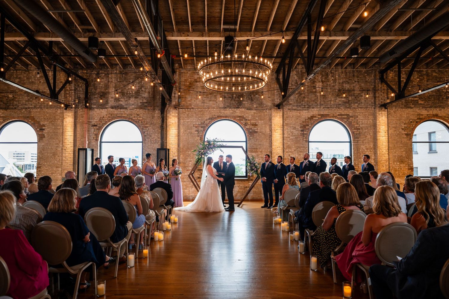 Elegant indoor wedding ceremony in a rustic brick venue with arched windows, chandelier lighting, and guests seated along a candlelit aisle.