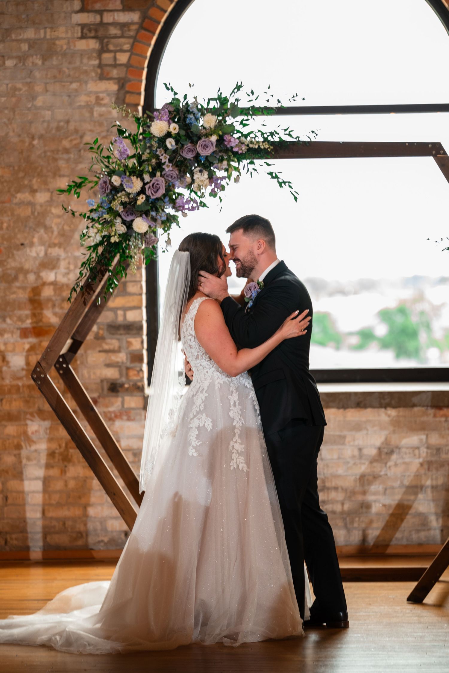 Bride and groom share a romantic moment under a geometric wooden arch adorned with purple and white florals in a rustic brick venue.