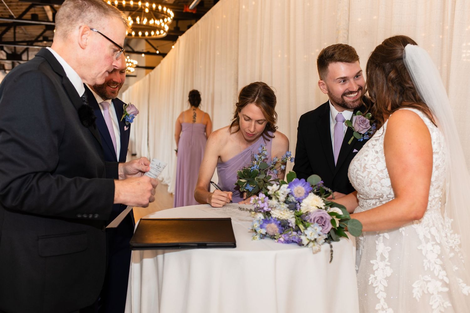 Bride and groom signing wedding documents with officiant and bridesmaid at elegant indoor ceremony.