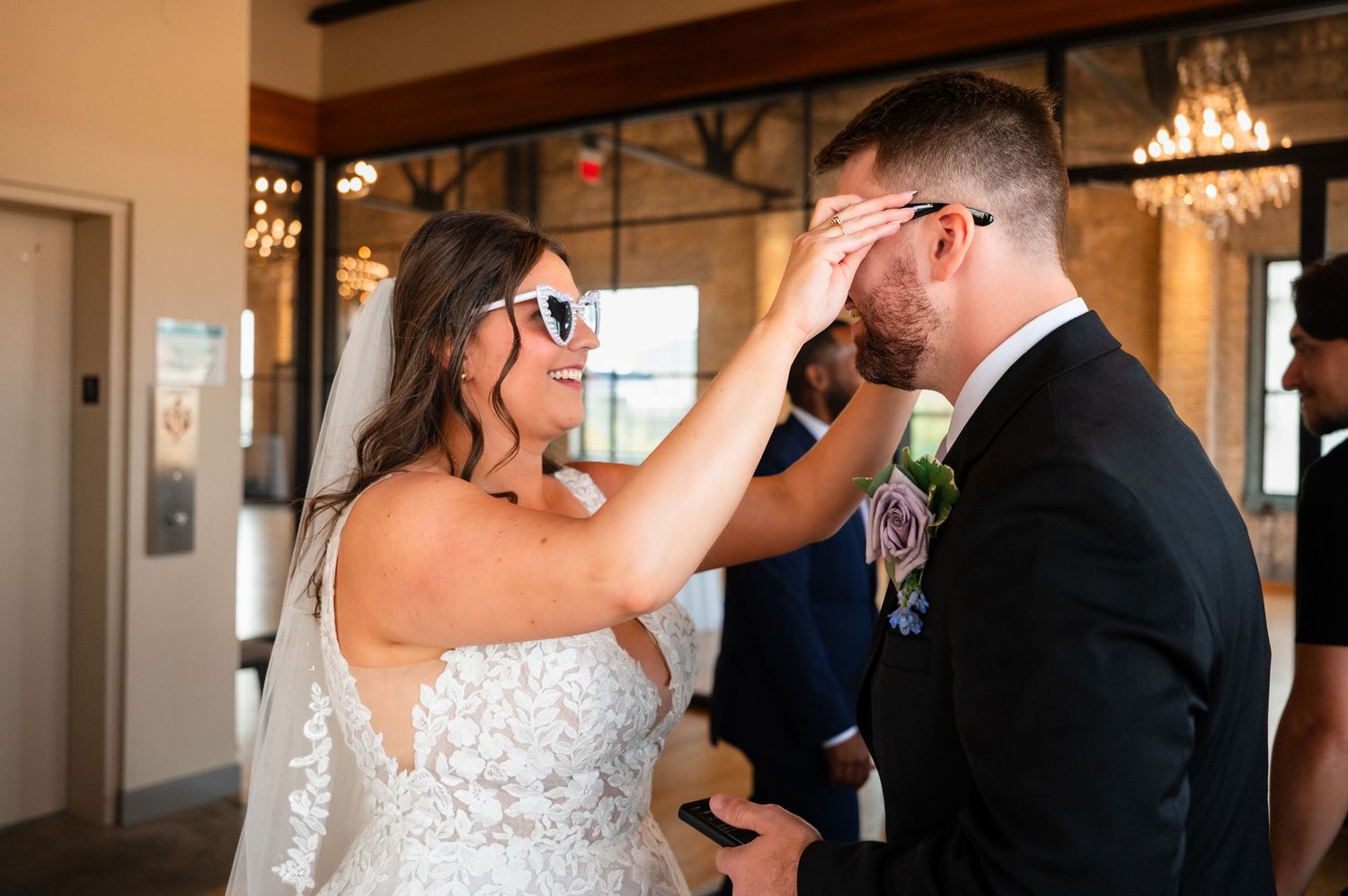Bride placing white sunglasses on groom during wedding ceremony in elegant venue with chandeliers.