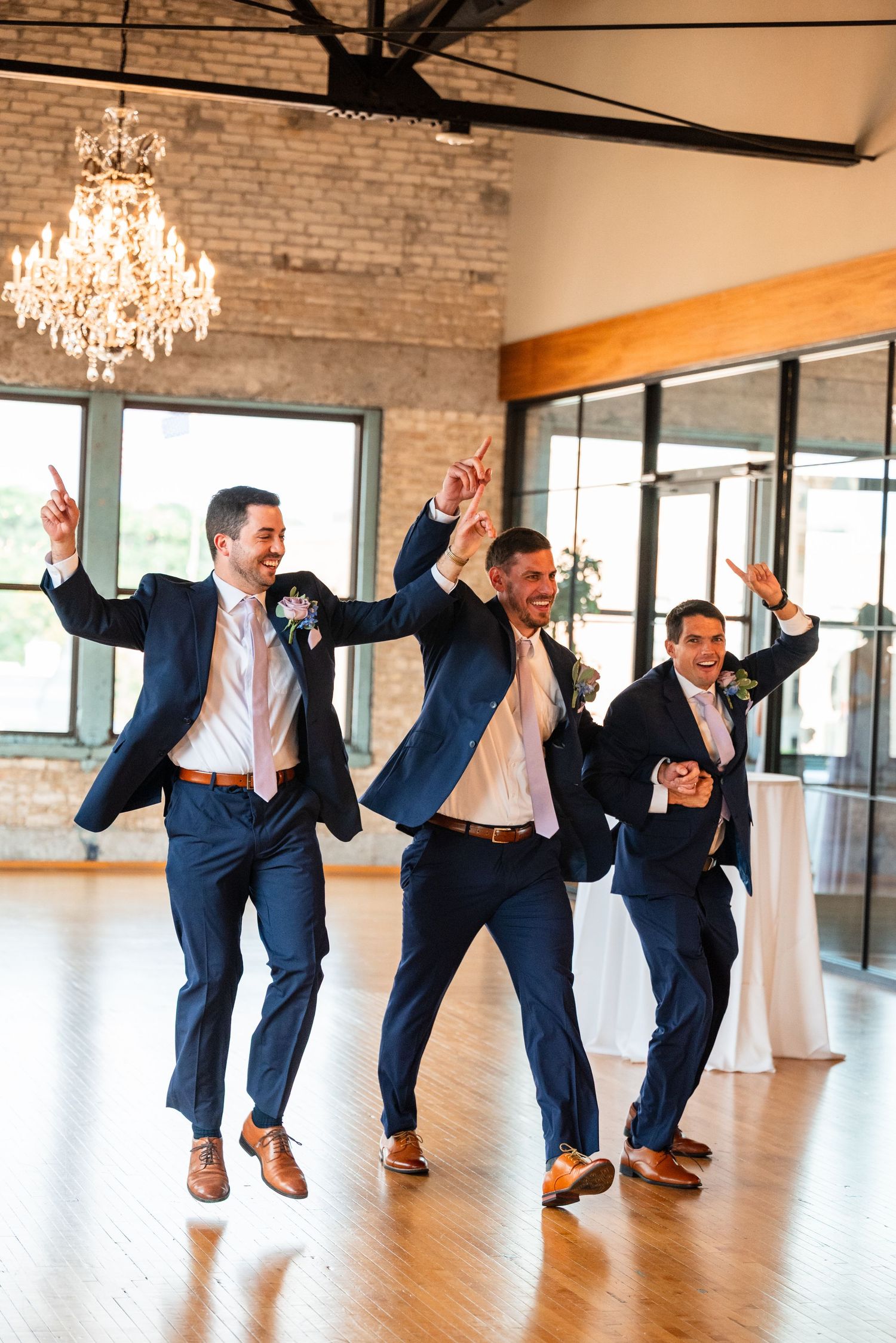 Three groomsmen in navy suits dancing joyfully at a wedding reception in an elegant venue with a chandelier.
