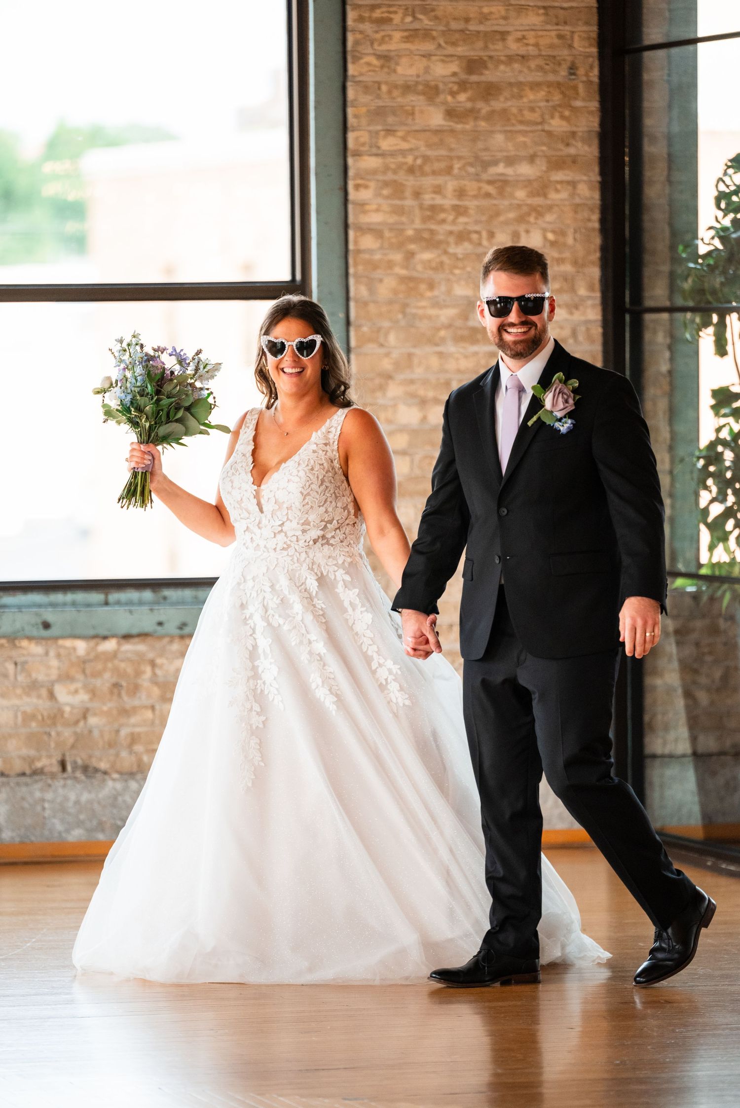 Bride in white lace gown and groom in black suit wearing fun sunglasses, holding hands and bouquet at wedding venue.
