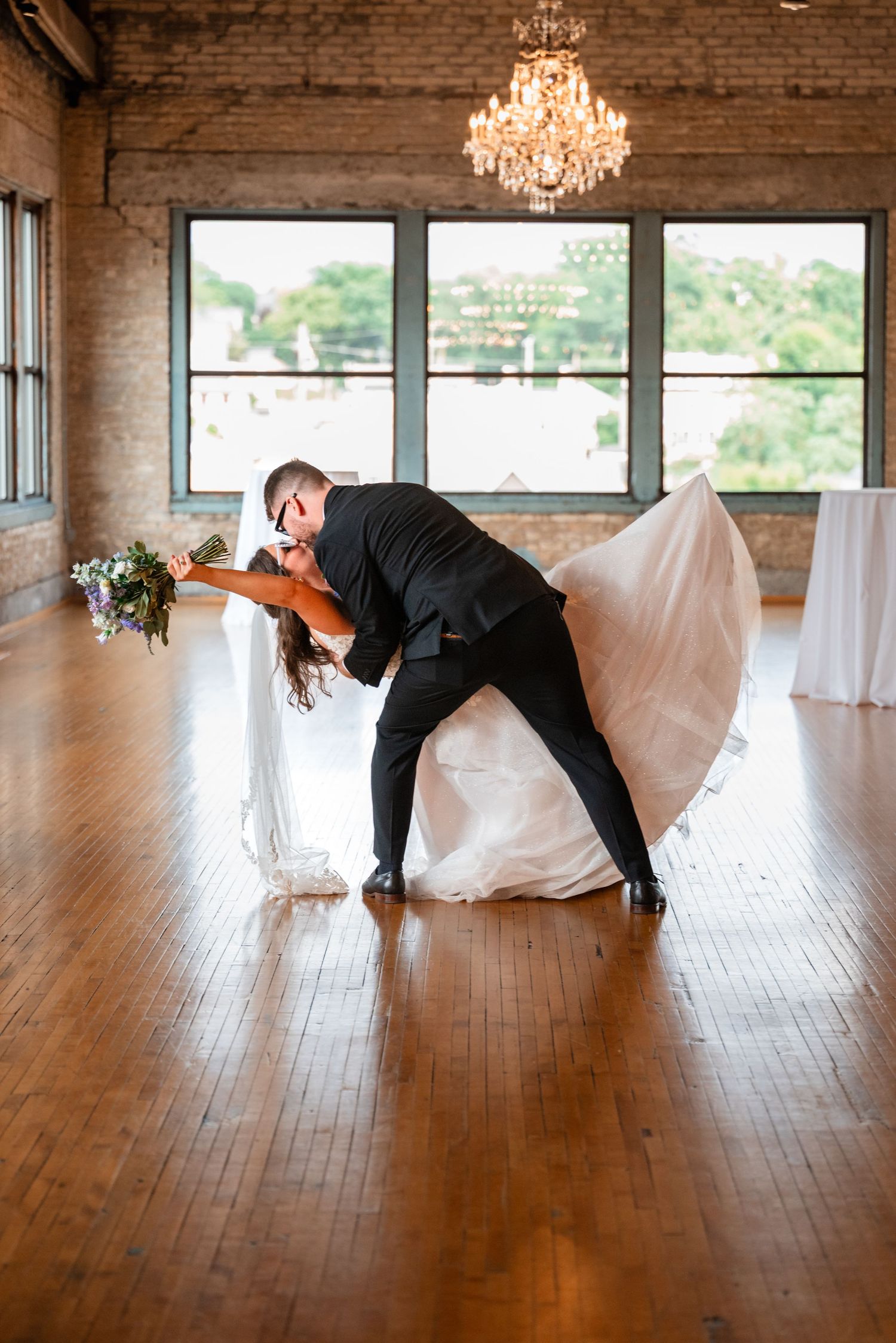 Bride and groom share a romantic dip kiss on a hardwood dance floor beneath a chandelier in a rustic brick venue.