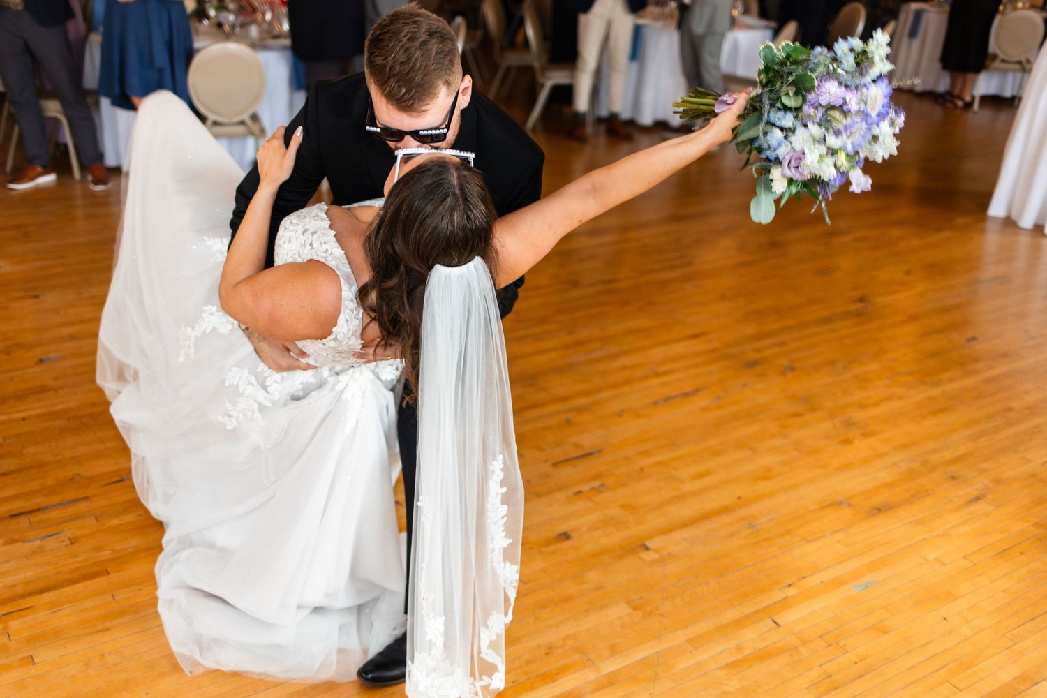 Groom dips bride on dance floor as she raises floral bouquet during romantic first dance at wedding reception.