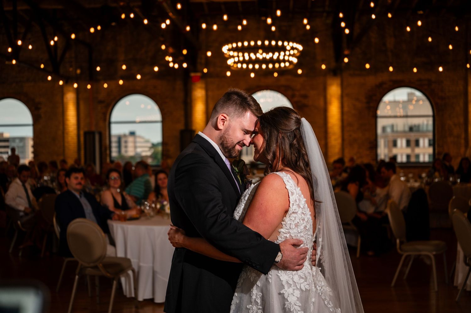 Bride and groom share intimate first dance under warm string lights in elegant brick wedding venue.