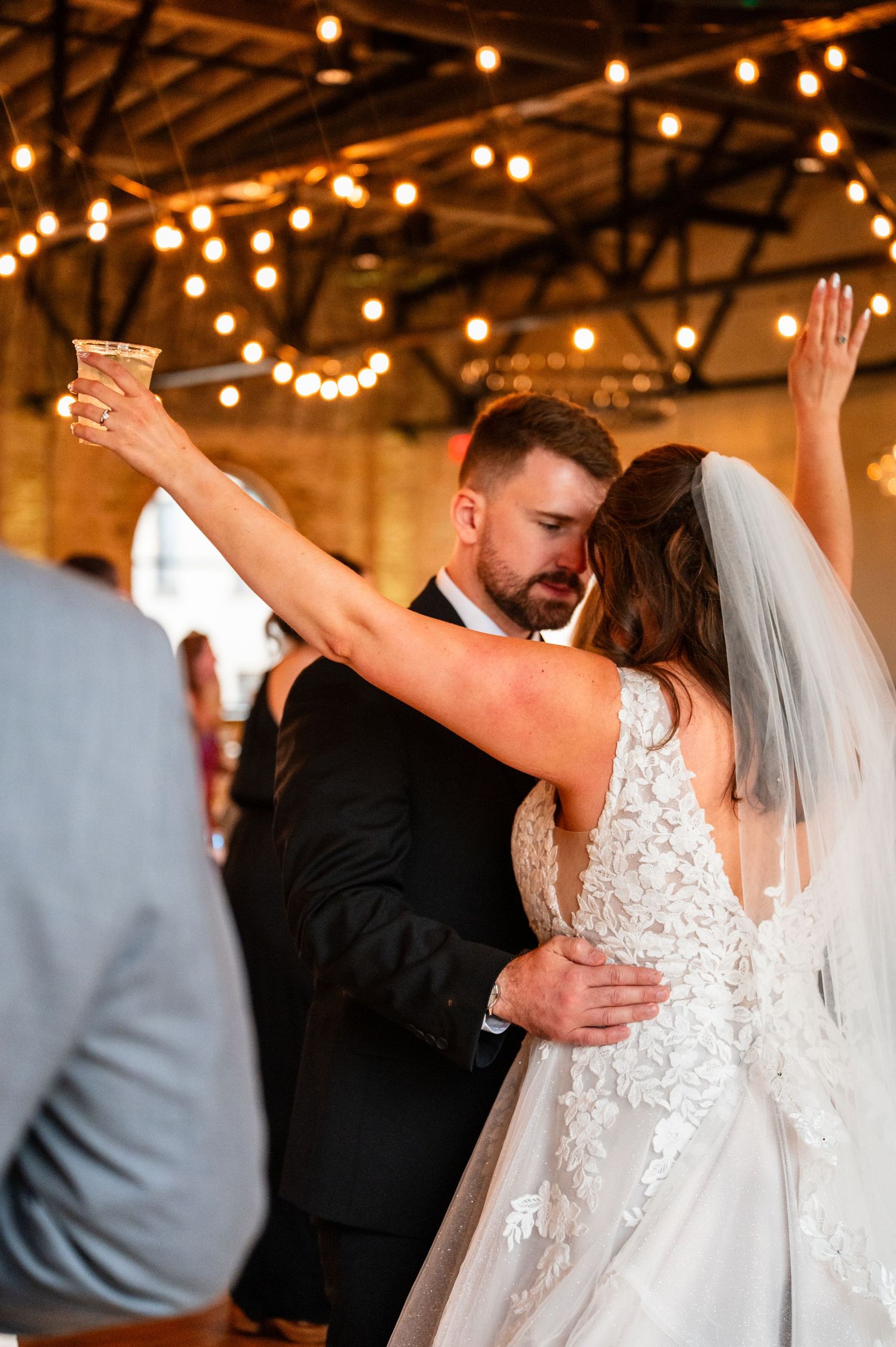 Bride and groom share an emotional first dance, bride raising a glass under warm string lights at wedding reception.