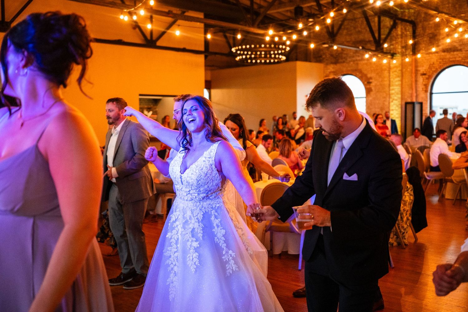 Bride in lace gown laughs joyfully while dancing with groom in black suit at elegant wedding reception.