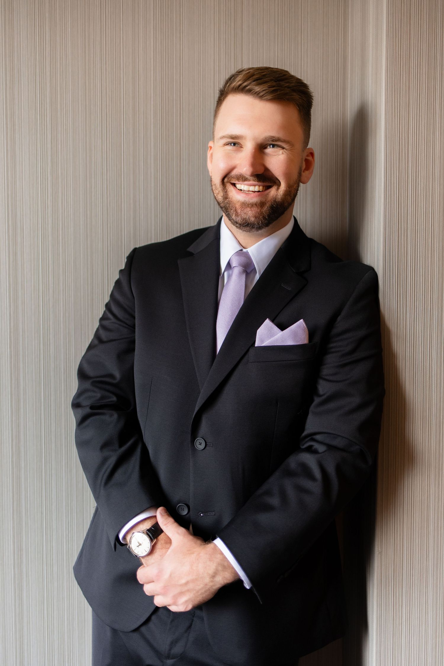 Smiling man in black suit with lavender tie and pocket square, wearing a watch, leaning against a wall.