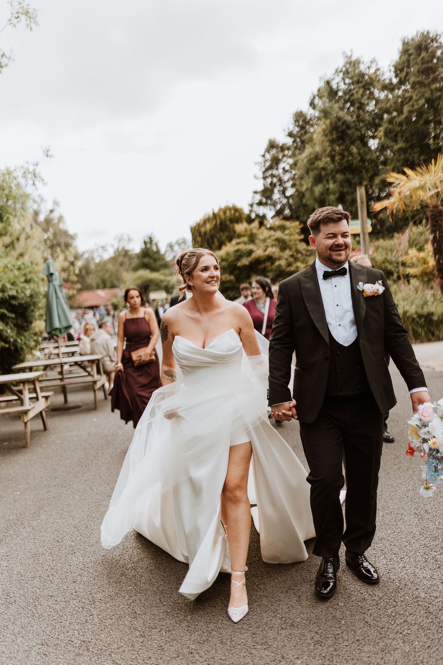 Smiling bride in white gown and groom in black tuxedo walk hand in hand at outdoor wedding celebration.