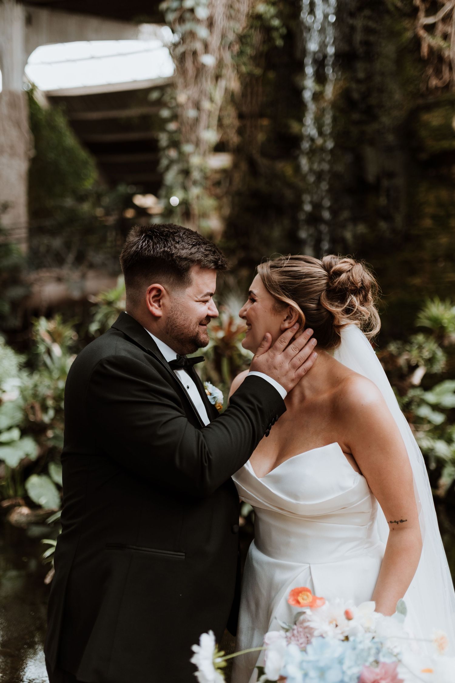 Groom tenderly holds bride's face in lush garden setting, both smiling, bride holding pastel bouquet in elegant white gown.