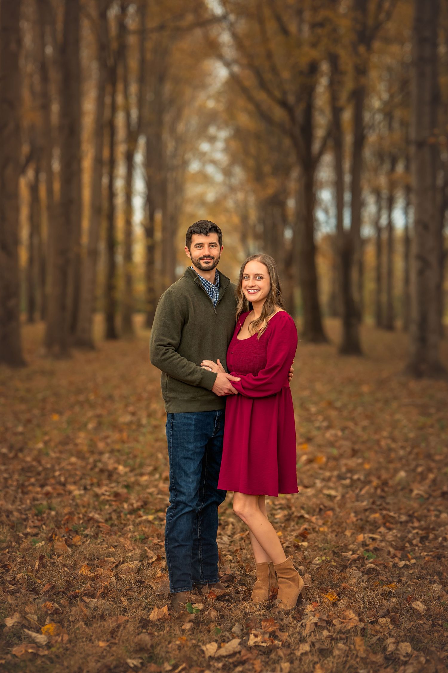 Couple posing together in autumn forest, woman in red dress and man in green sweater, surrounded by fall foliage.