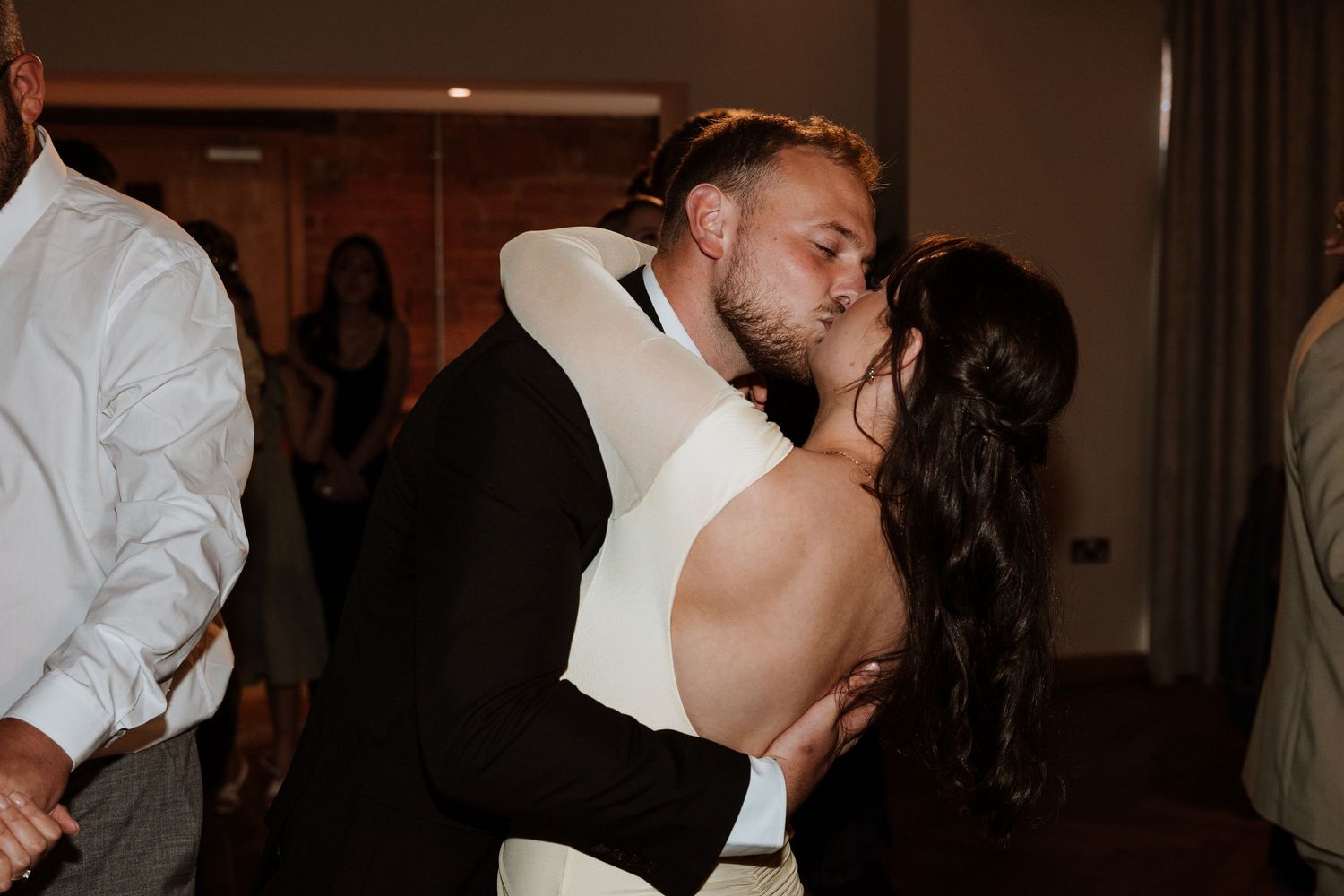 A couple shares a romantic first dance kiss at their wedding reception, bride in backless white gown.