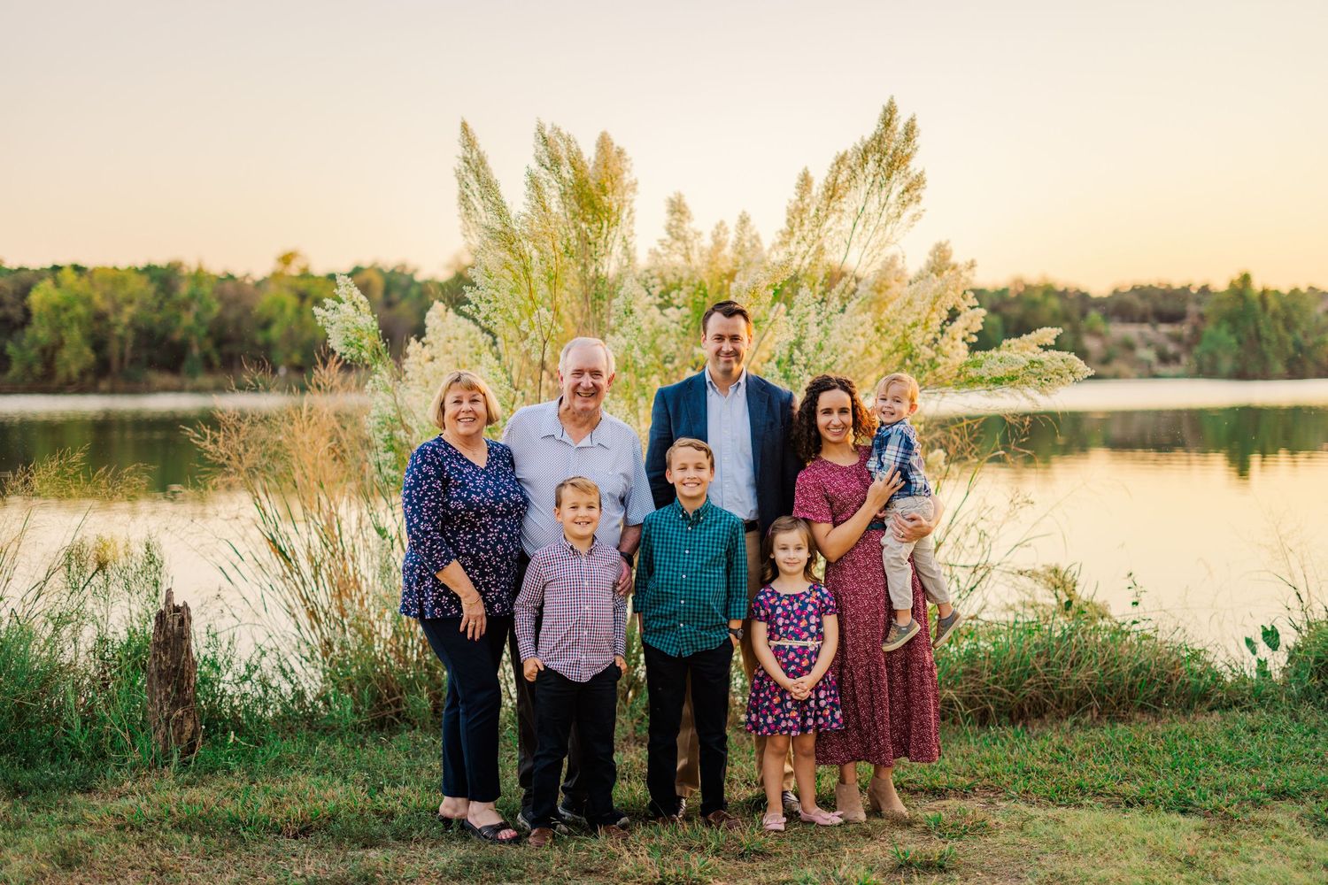Family portrait of grandparents, parents, and four children smiling near a serene lake at golden hour sunset.