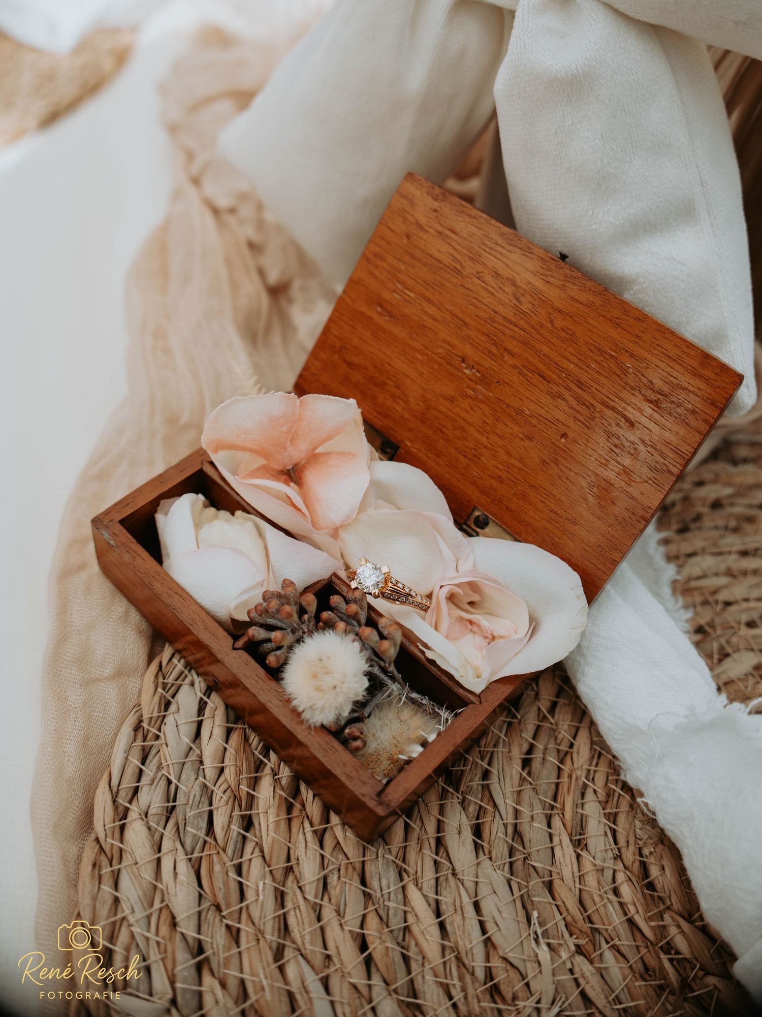 Open wooden ring box with dried flowers and wedding rings, styled with woven basket and soft fabric backdrop.