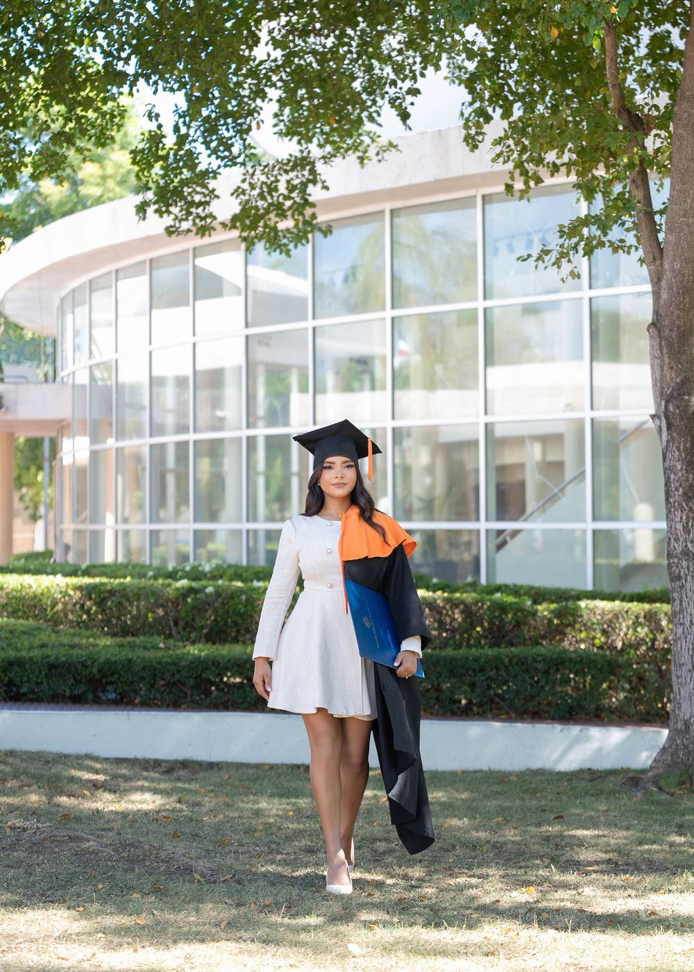 Young woman in white dress and graduation cap holding black and orange gown outdoors near modern building.