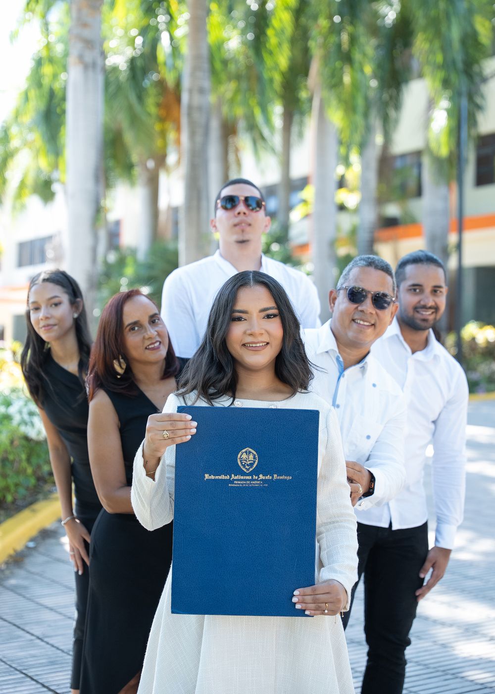 Young woman proudly holds blue university diploma folder surrounded by smiling family outdoors near palm trees.