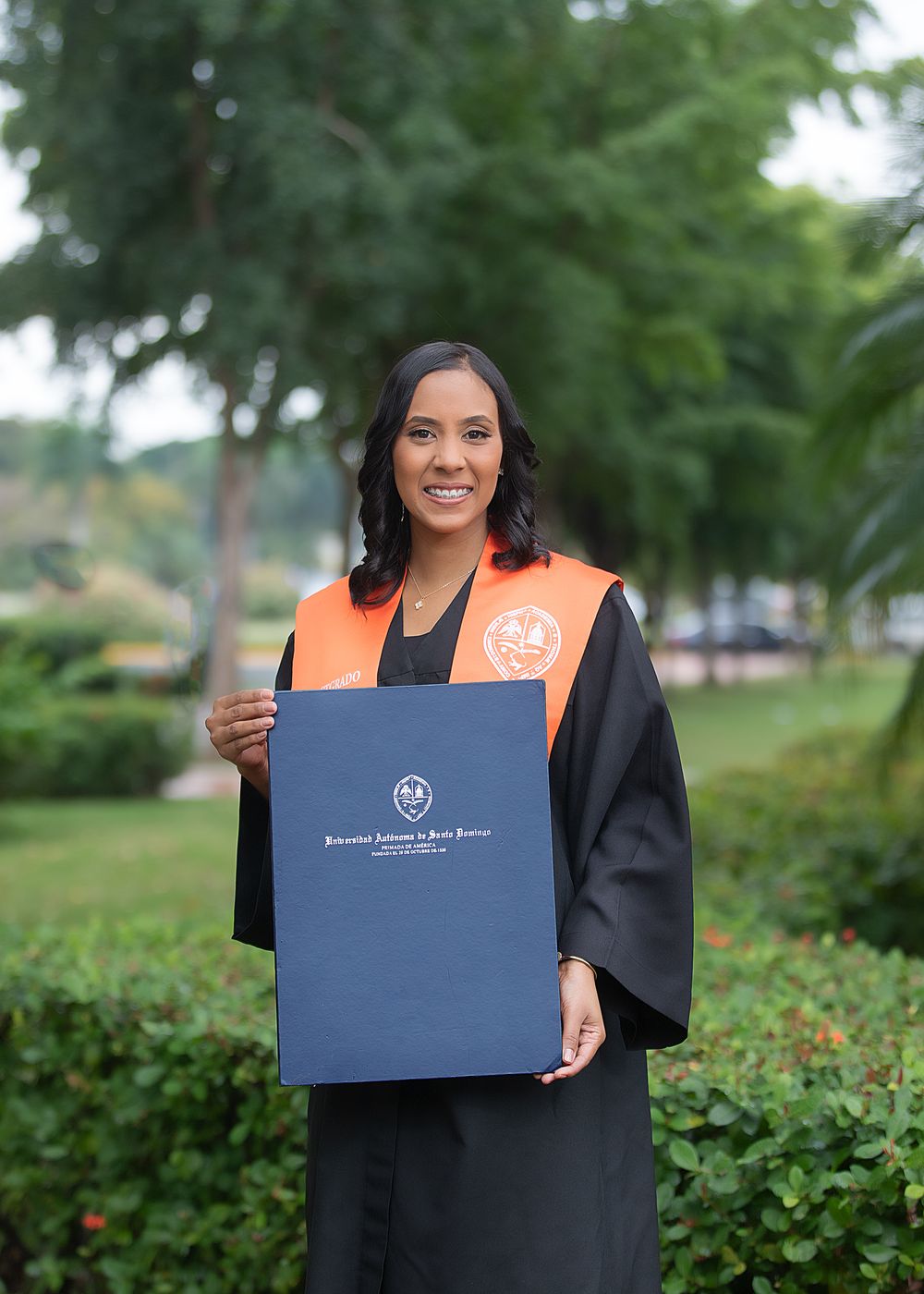 A smiling woman in black graduation gown and orange sash holds a diploma folder outdoors among green trees.