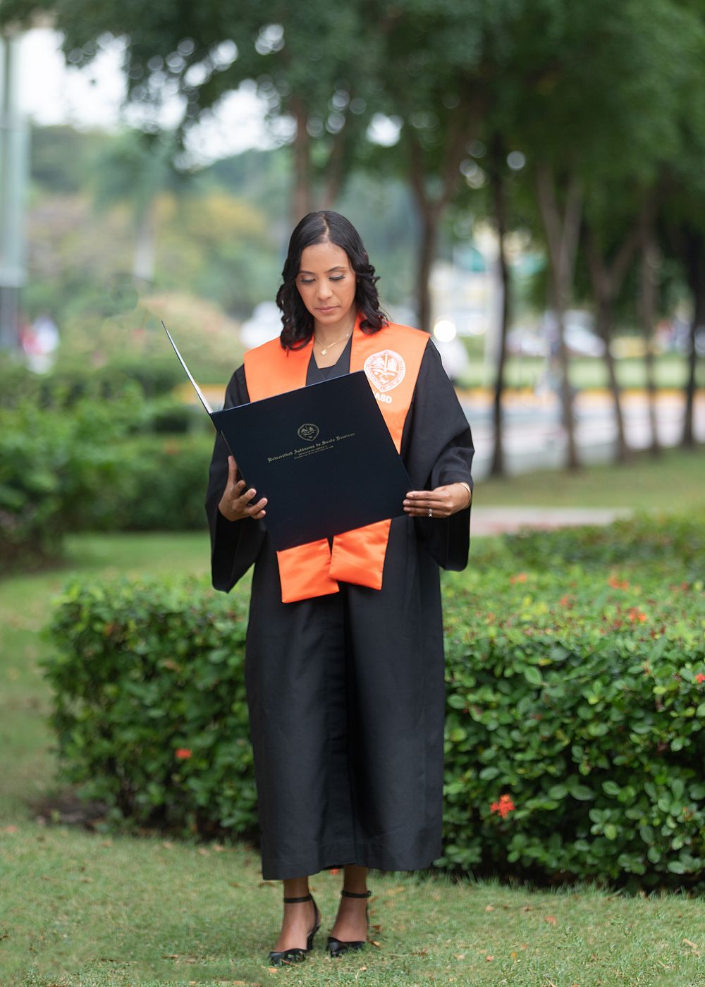 A woman in black and orange graduation gown holds her diploma in a lush green park setting.