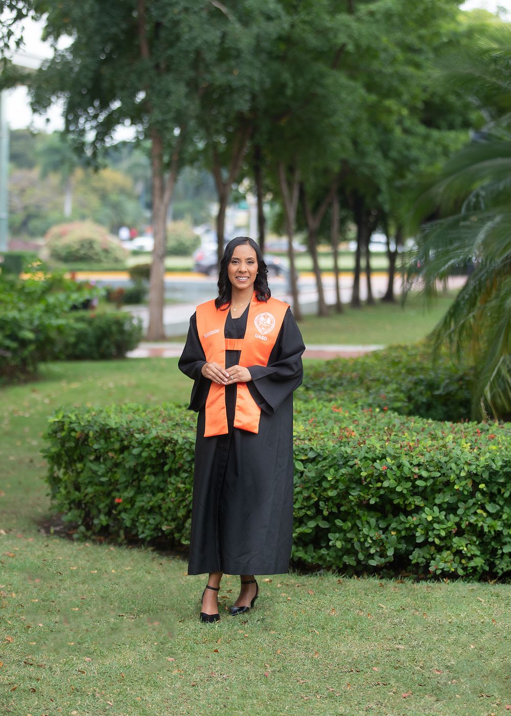 Woman in black graduation gown with orange stole stands on green campus lawn surrounded by tropical trees.