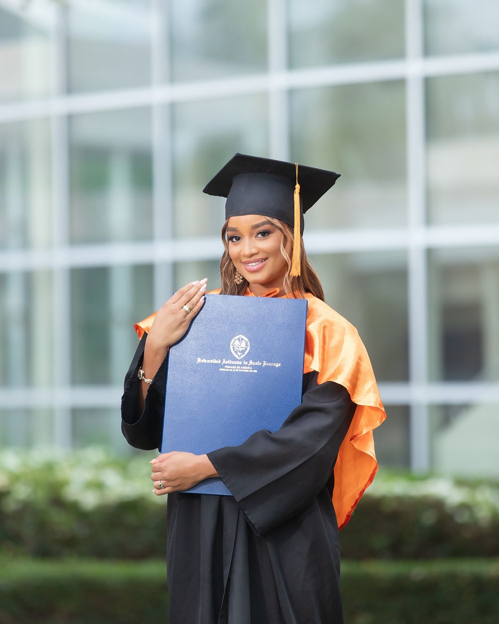 A smiling woman in black graduation gown and cap with orange sash holds a blue diploma outside a modern building.