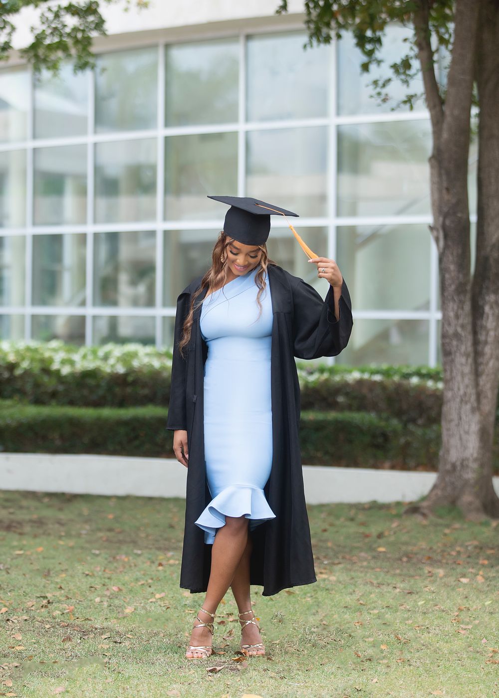 A woman in a light blue dress and black graduation cap and gown holds her tassel outdoors on a sunny day.