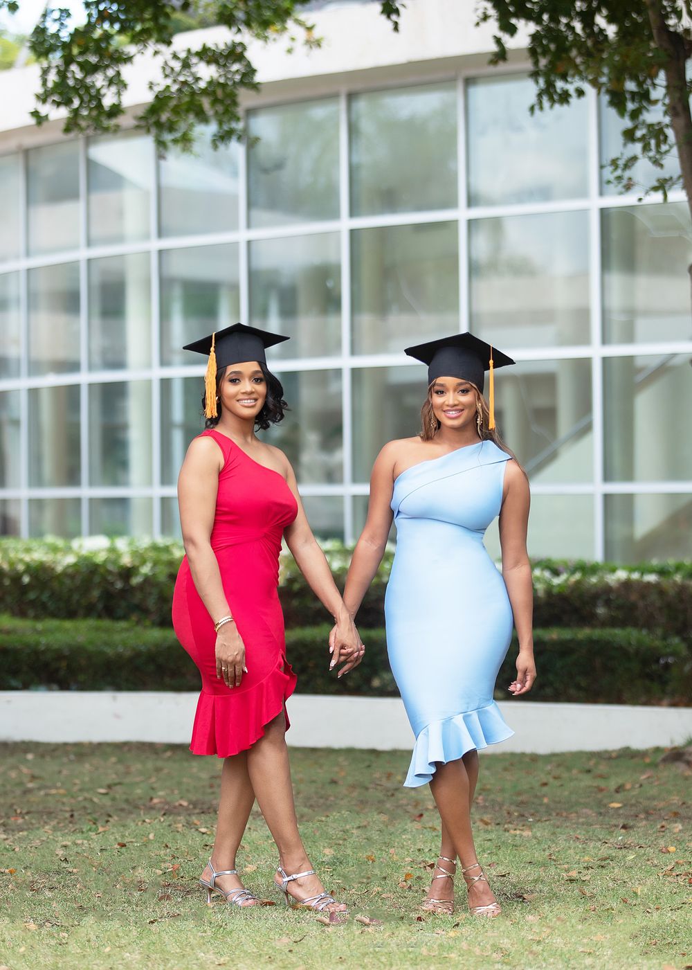 Two women in graduation caps, one in red dress and one in light blue dress, holding hands outdoors.